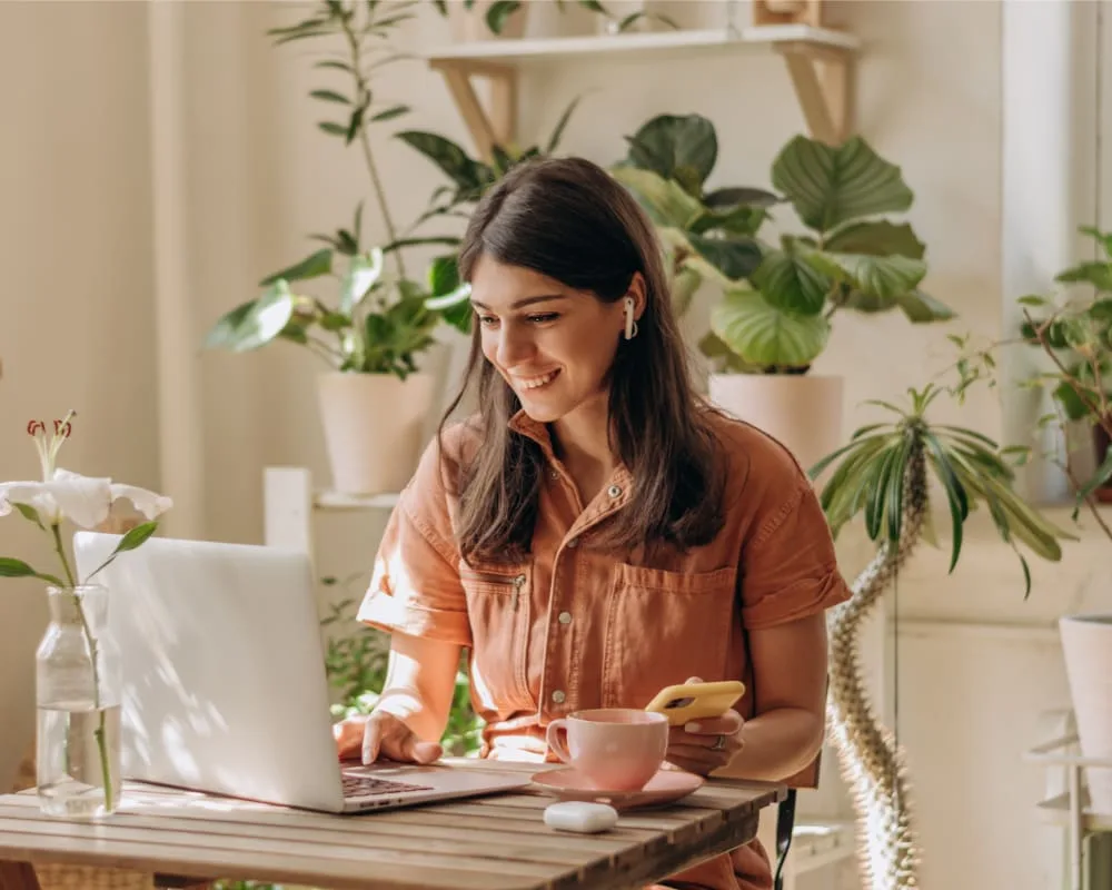 Woman working on a laptop in a plant-filled setting.