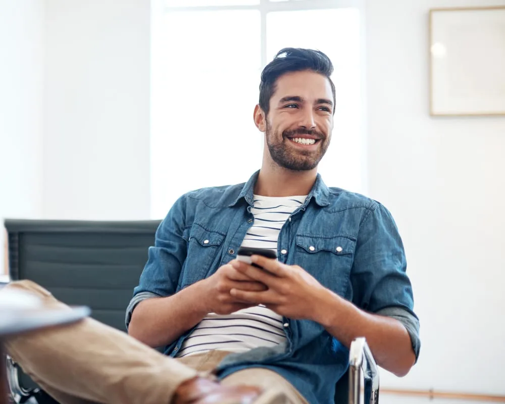 A smiling man in a denim jacket holding a smartphone.