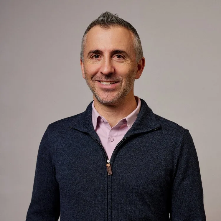 A man in a navy sweater and pink shirt smiling.