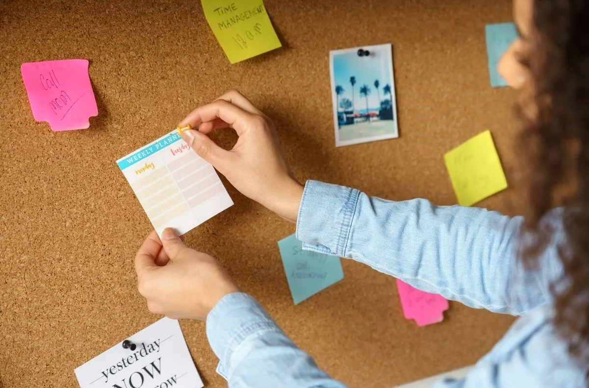 A person arranging a weekly planner on a corkboard.