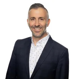 A smiling man in a dark blazer and patterned shirt.