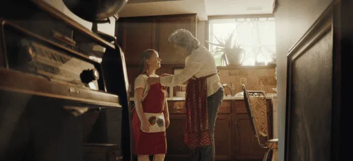 A grandmother adjusts a child's apron in a kitchen scene.