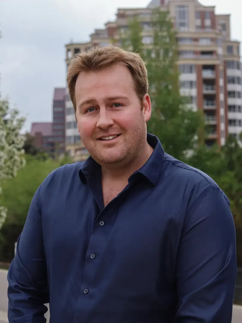 A man smiling outdoors with modern buildings behind him.