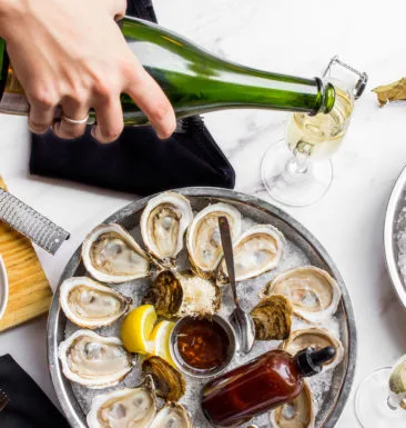 Hand pouring wine into a champagne flute next to oysters.
