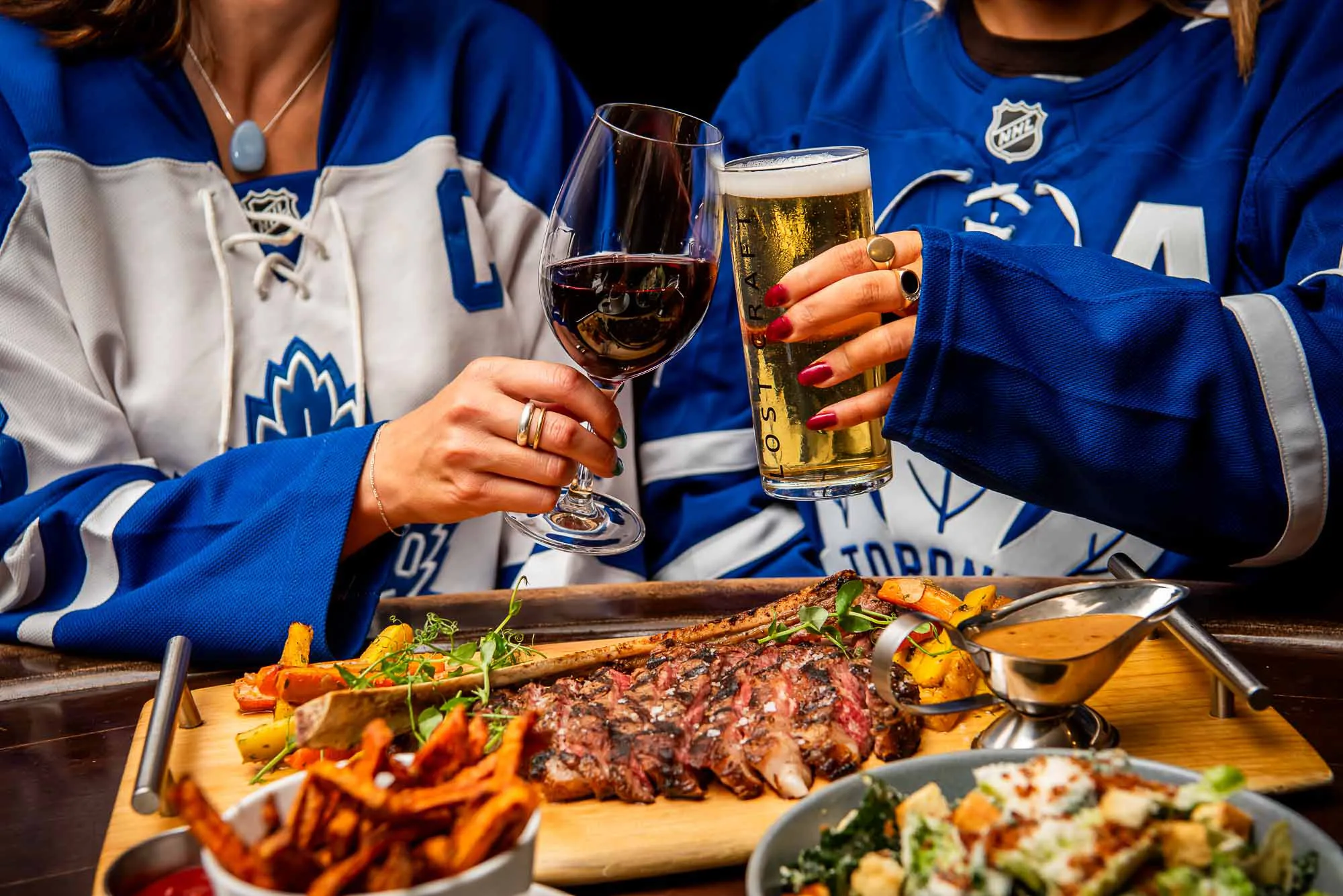 Two women toasting with drinks over a platter of food.
