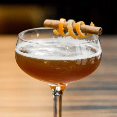A cocktail in a coupe glass garnished with cinnamon and orange peel.