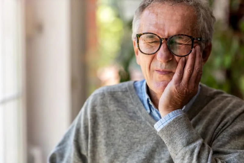 An older man with glasses looks pained while holding his cheek.