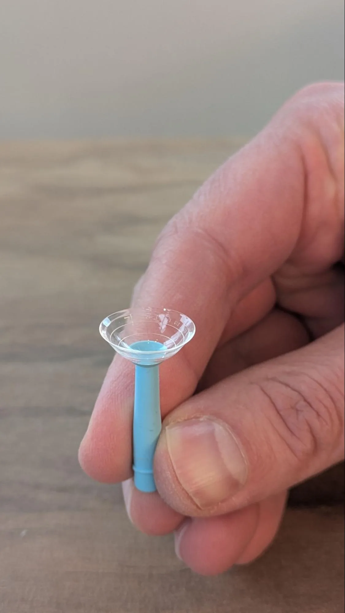 A hand holding a filled scleral contact lens on a blue plunger.