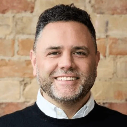 A smiling man with dark hair and a beard against a brick wall.