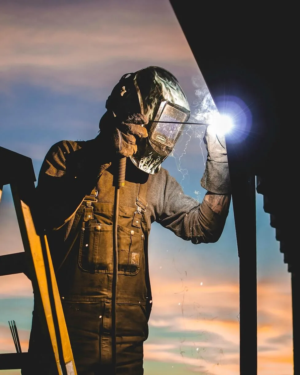 A welder working on metal with a colorful sky behind.