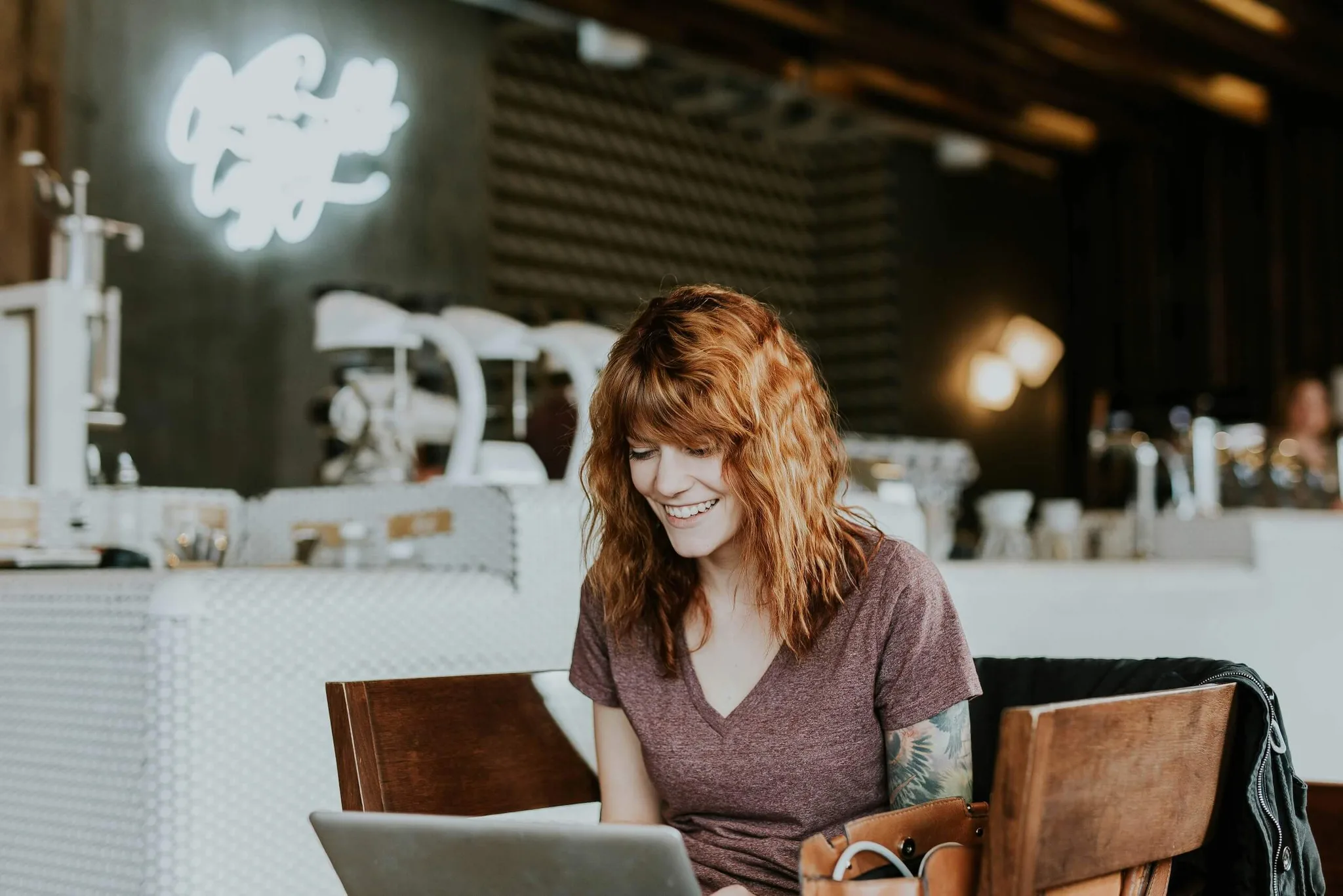 A woman with red hair smiles at her laptop in a cafe.