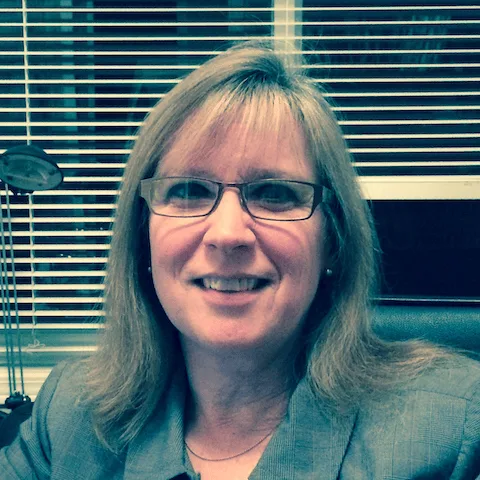 A smiling woman in a gray blazer in an office setting.