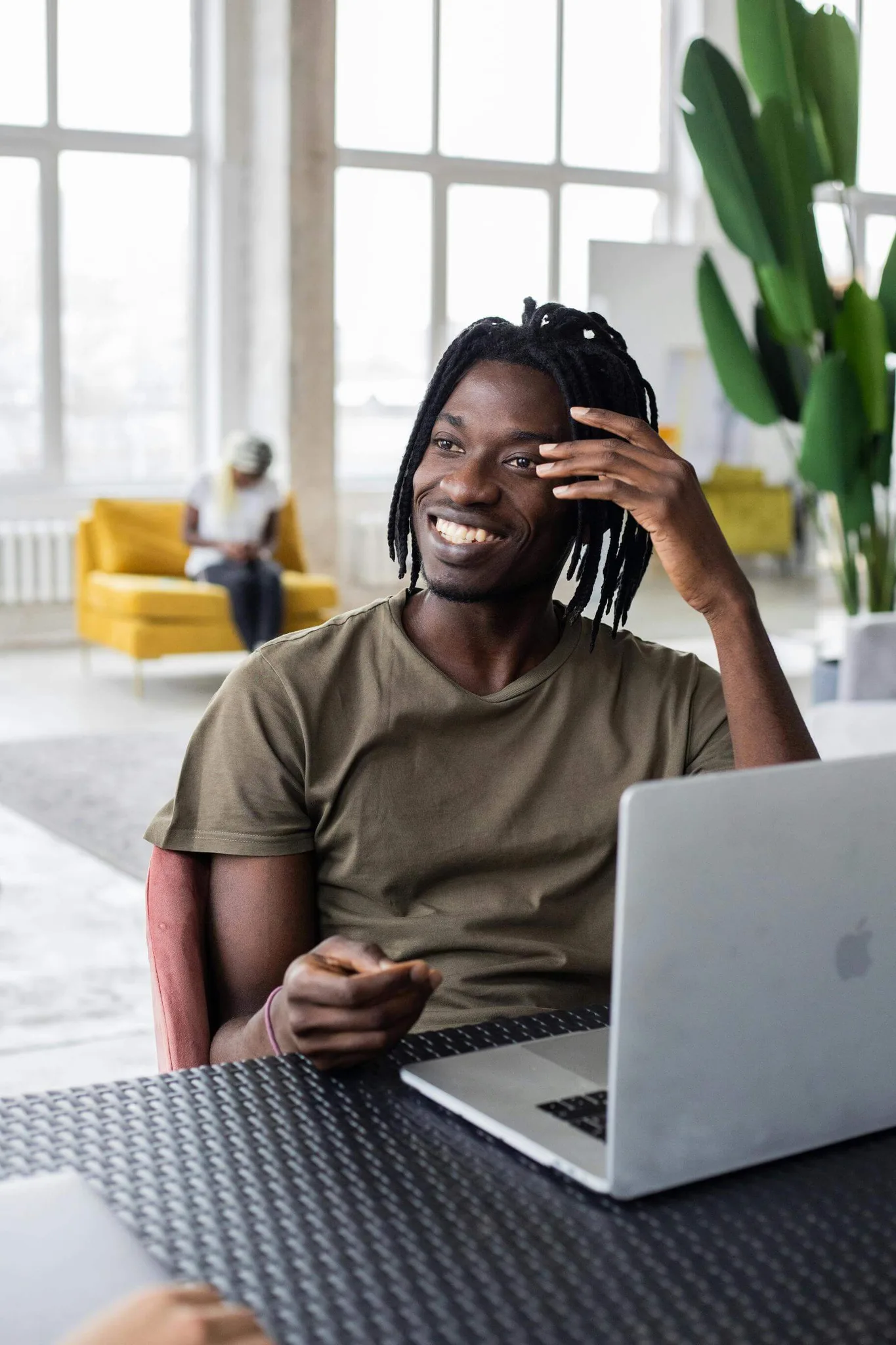 A smiling man with braided hair using a laptop in a bright workspace.
