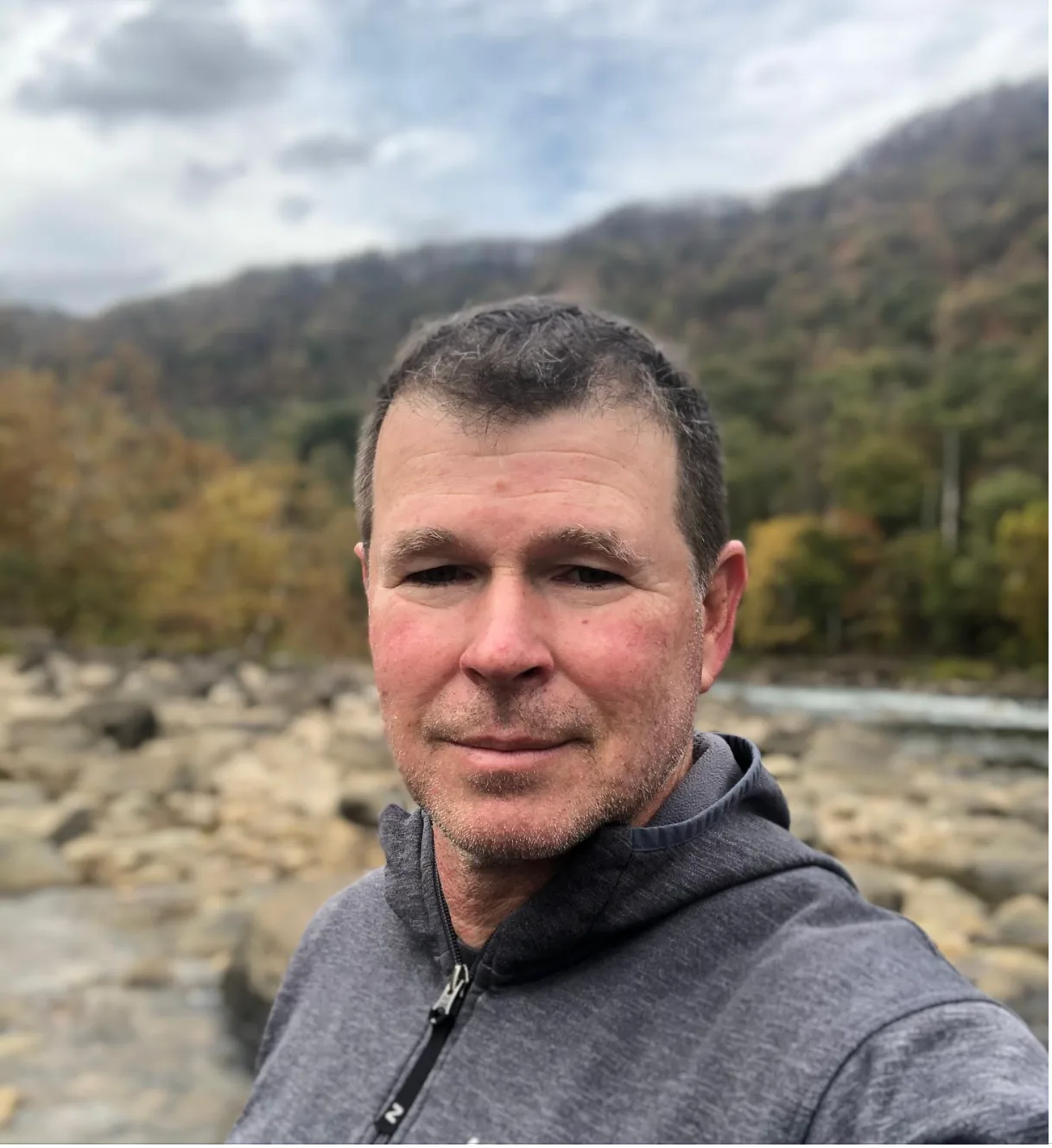 A man by a river with rocky terrain and trees in the background.