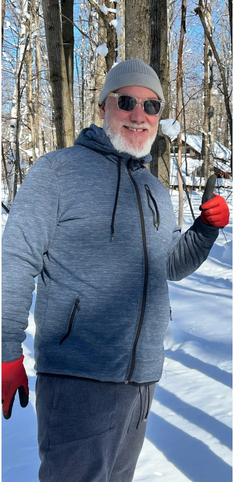 A smiling man in a snowy forest wearing a grey jacket and red gloves.