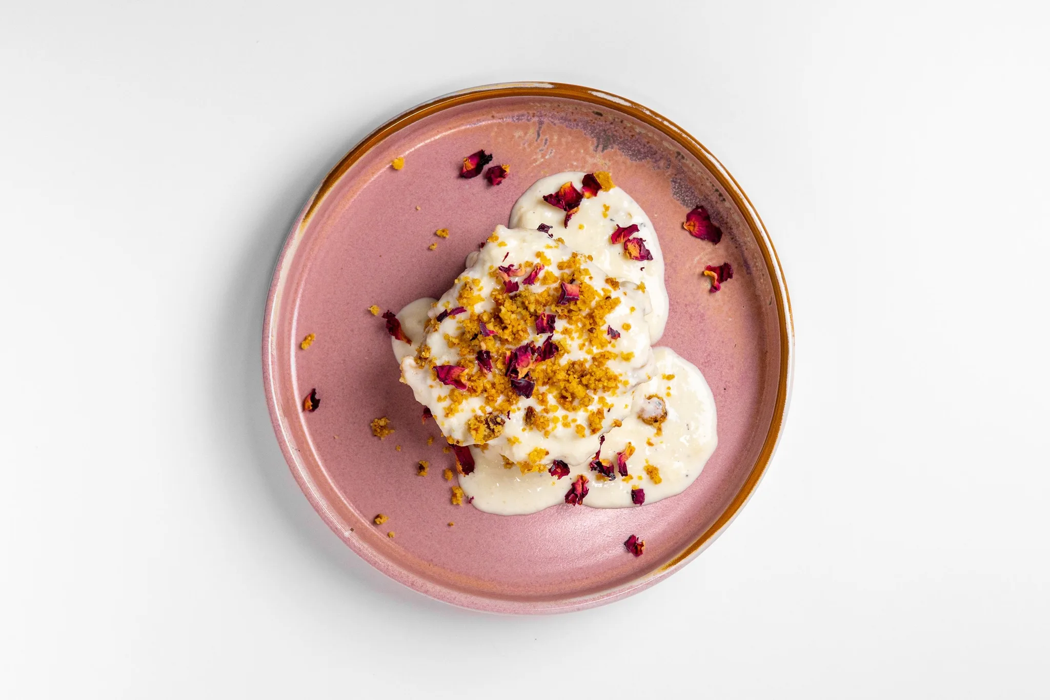 A creamy dessert with rose petals and crushed toppings on a pink plate.