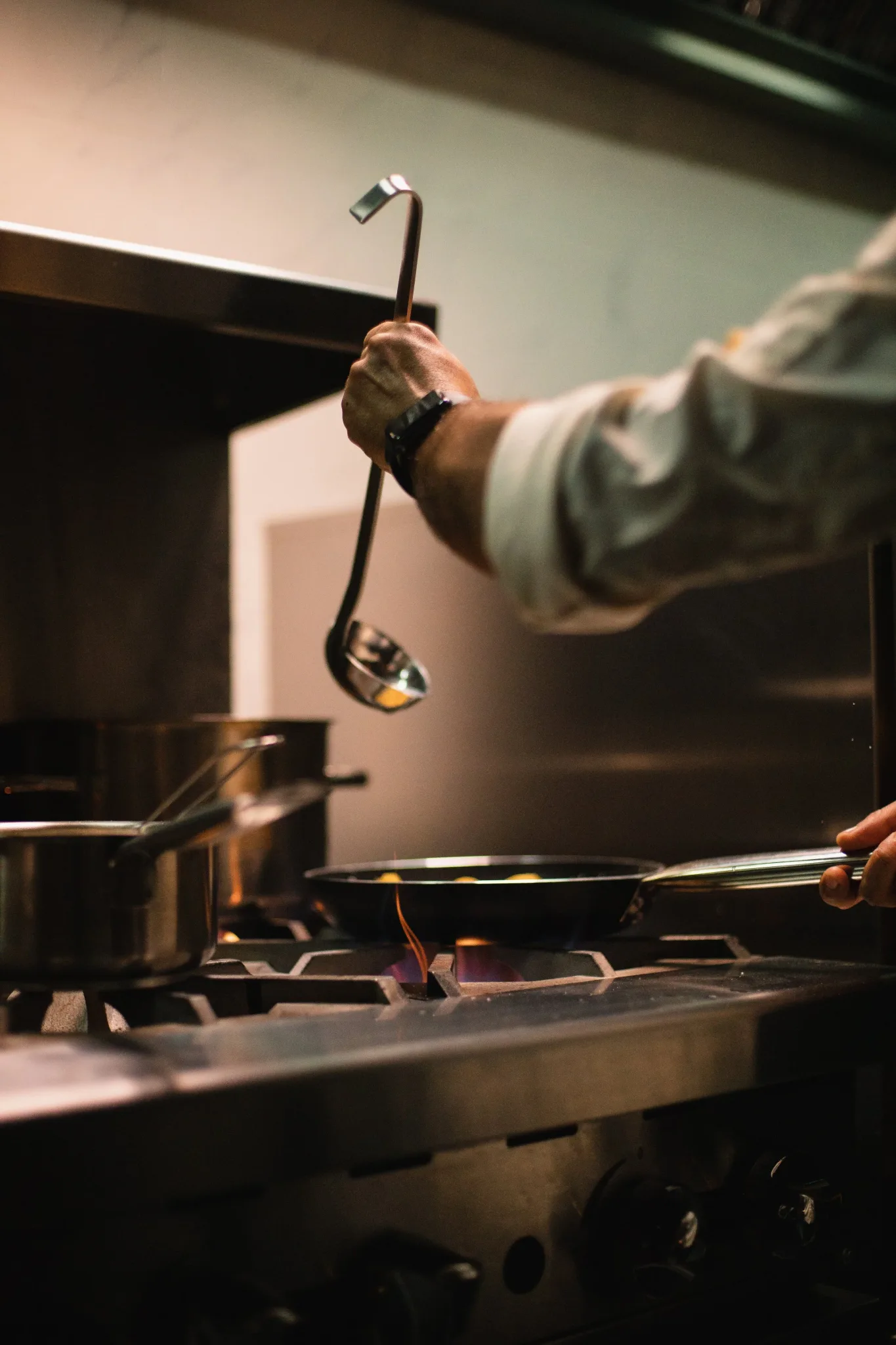A chef cooking in a modern kitchen with a ladle and frying pan.
