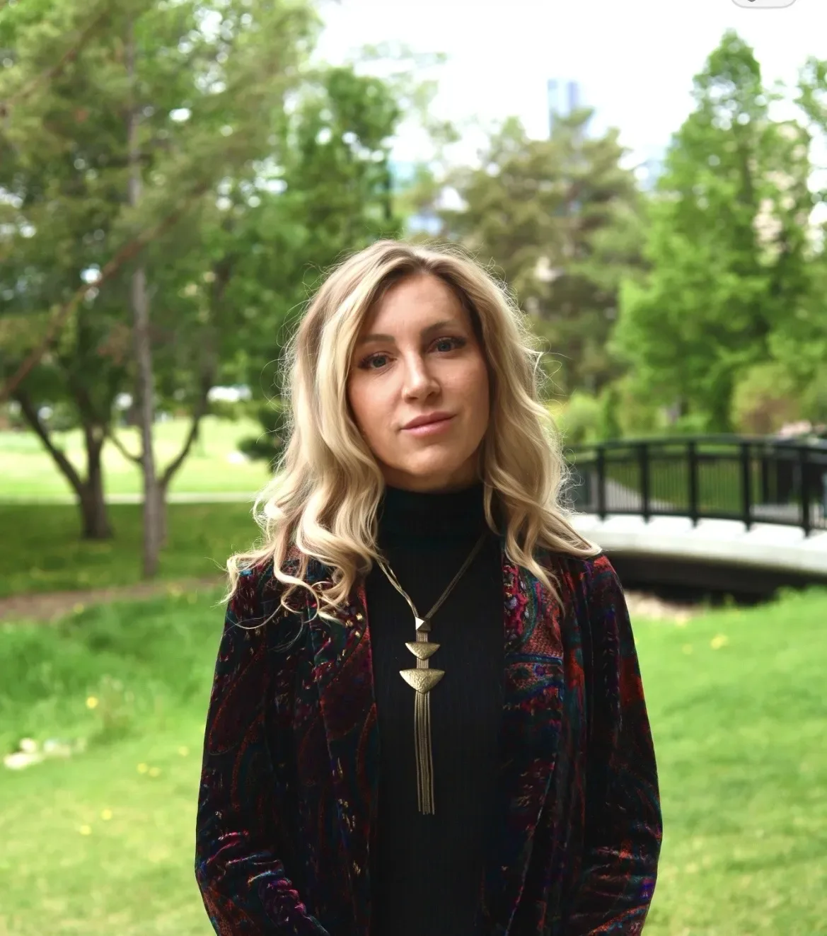 A woman in a park wearing a colorful jacket and necklace.