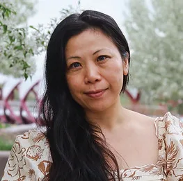 A woman posing outdoors with greenery in the background.