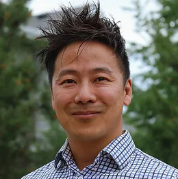 A smiling man outdoors with spiky hair in a checked shirt.