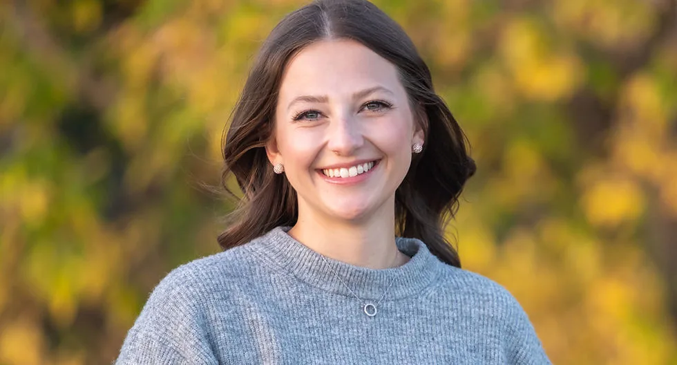 A woman smiling in a gray sweater with autumn leaves behind her.