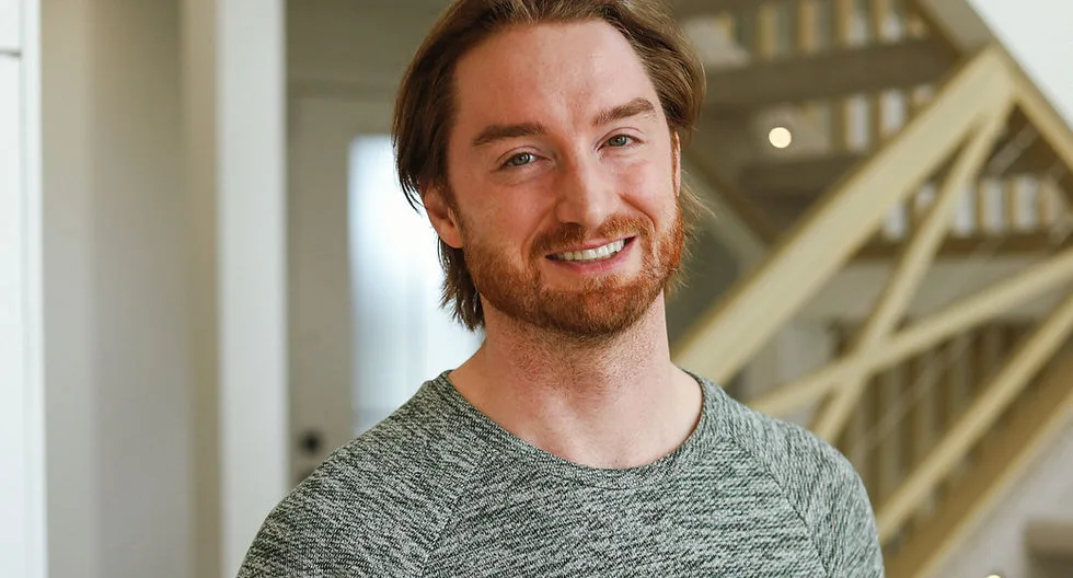 A smiling man in a grey athletic shirt standing indoors.
