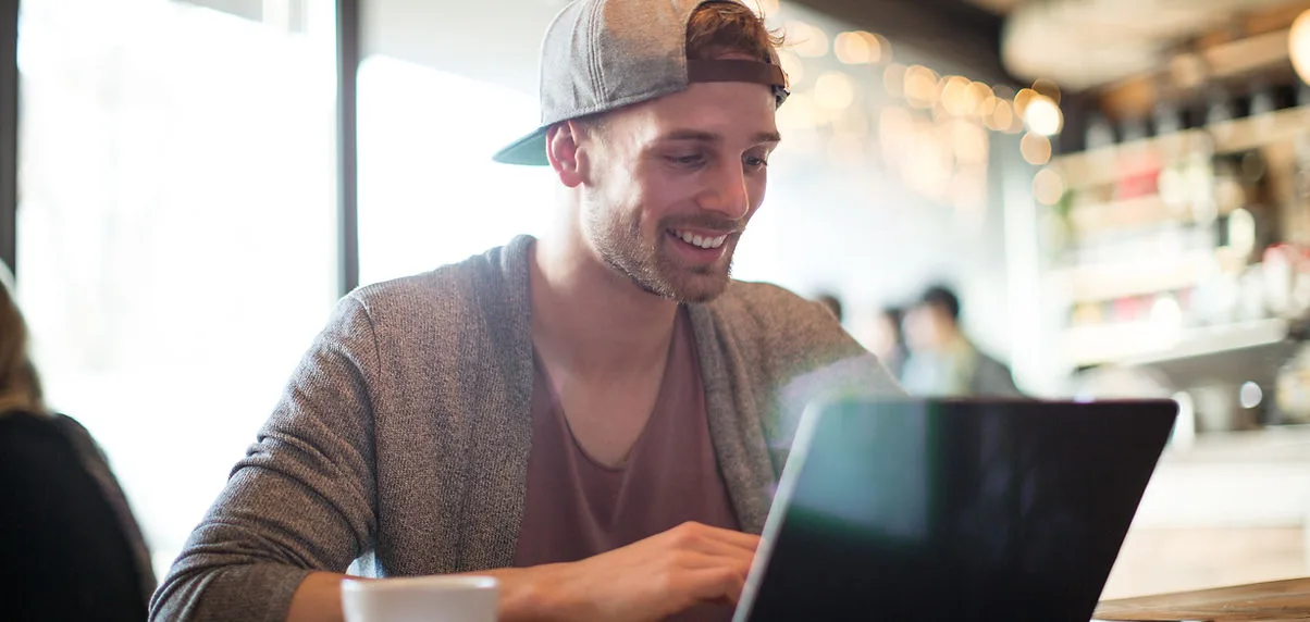 A man smiling while working on a laptop in a café.