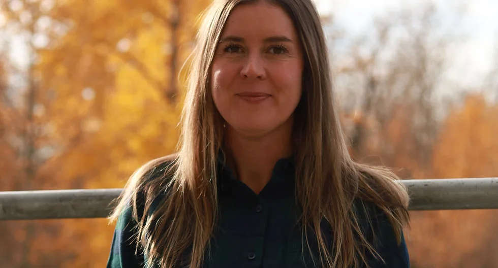 A woman outdoors in a plaid shirt with autumn trees behind her.