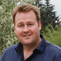 Man smiling in an outdoor setting surrounded by greenery.