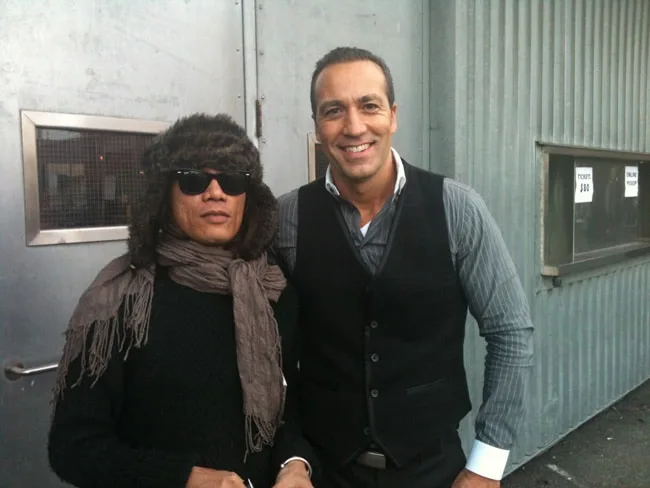 Two men posing outside a building, one in sunglasses and a hat.
