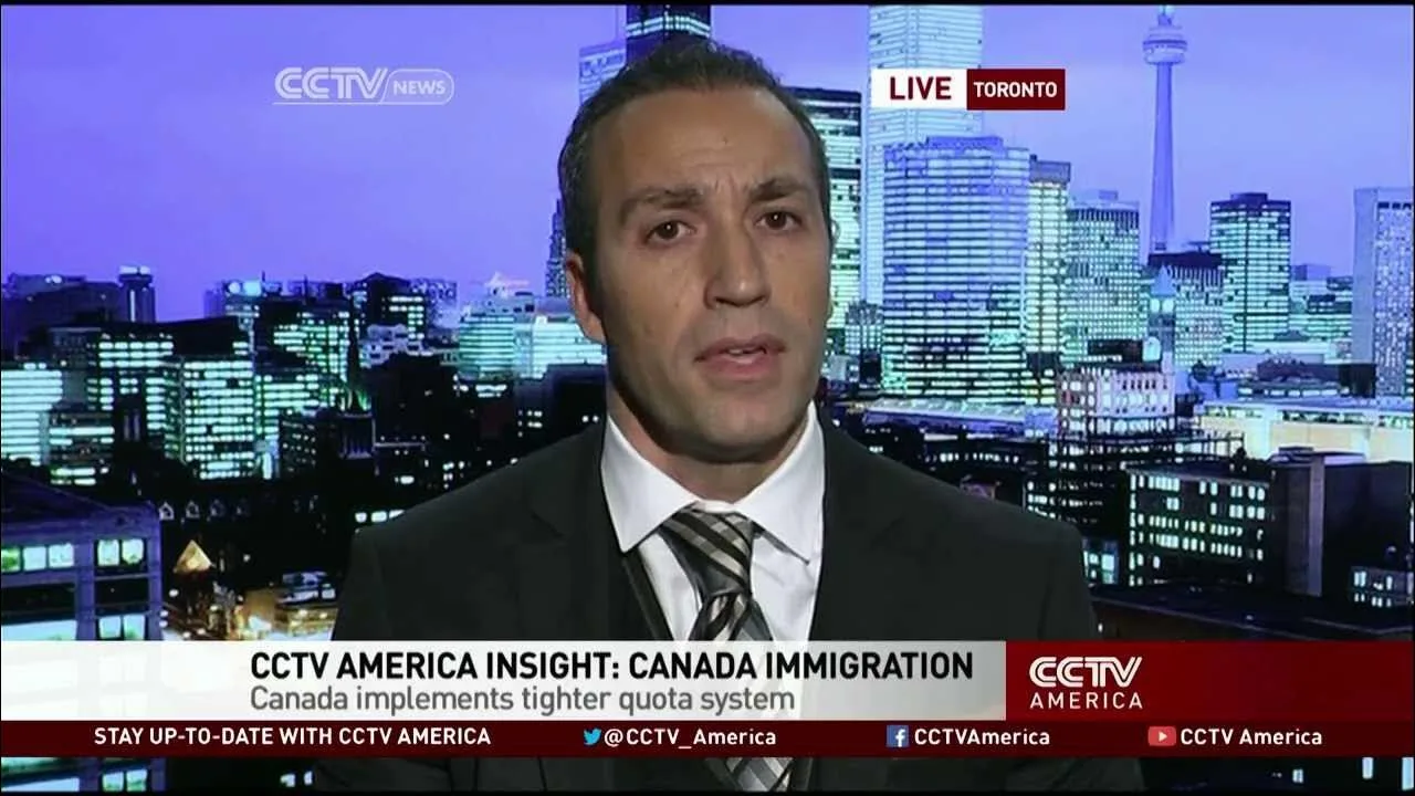 Immigration lawyer Henry Moyal speaking on a broadcast in front of Toronto skyline.