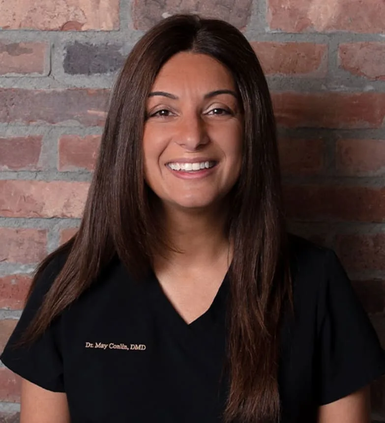 A smiling dentist in black scrubs in front of a brick wall.