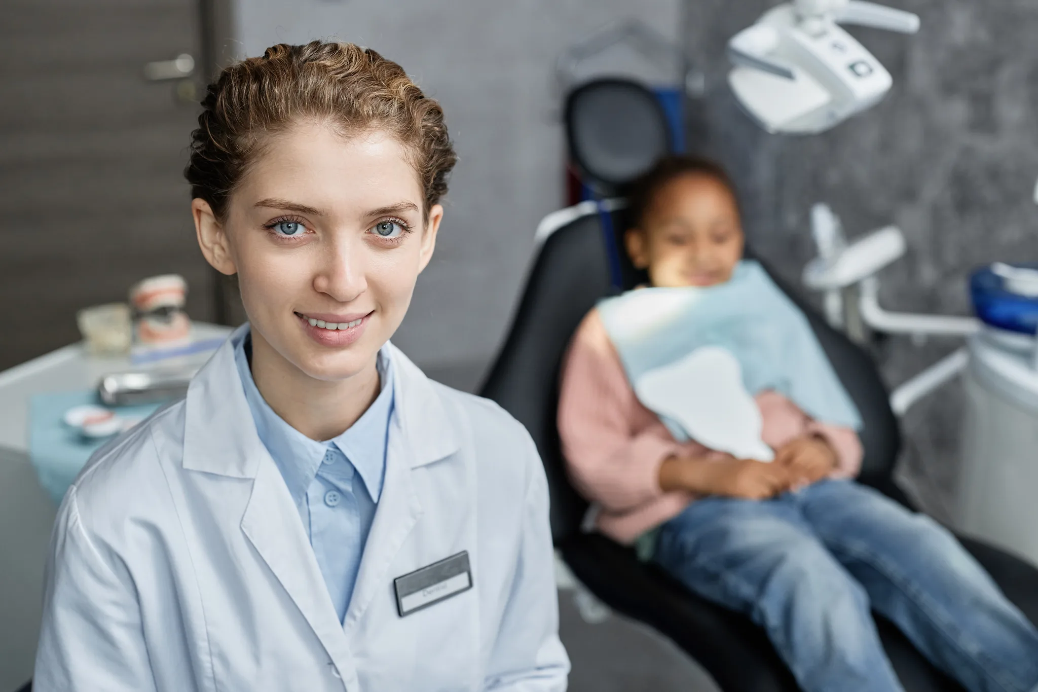 A young female dentist smiling in a clinic with a child patient in the background.