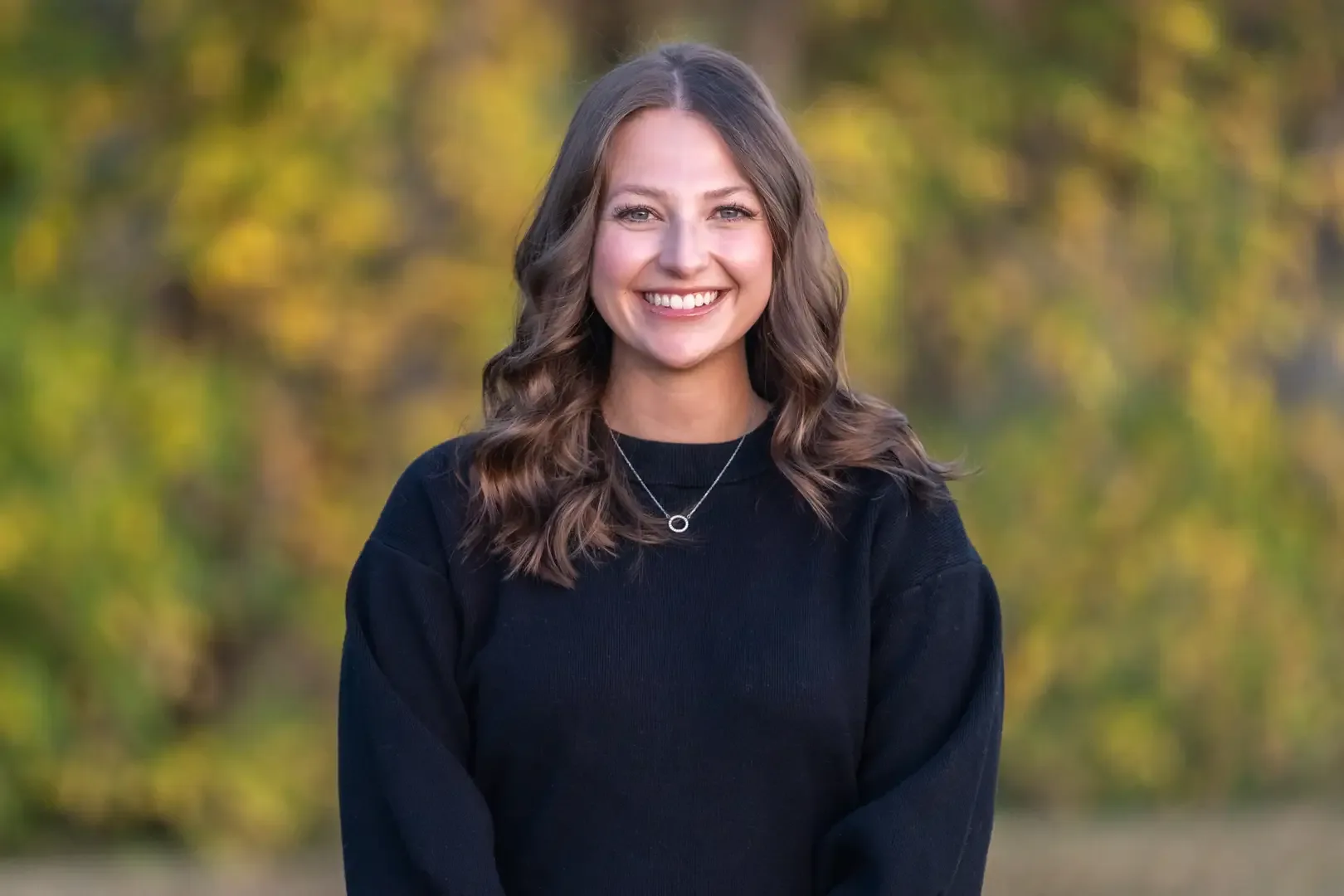 Smiling woman in black sweater with autumn foliage background.