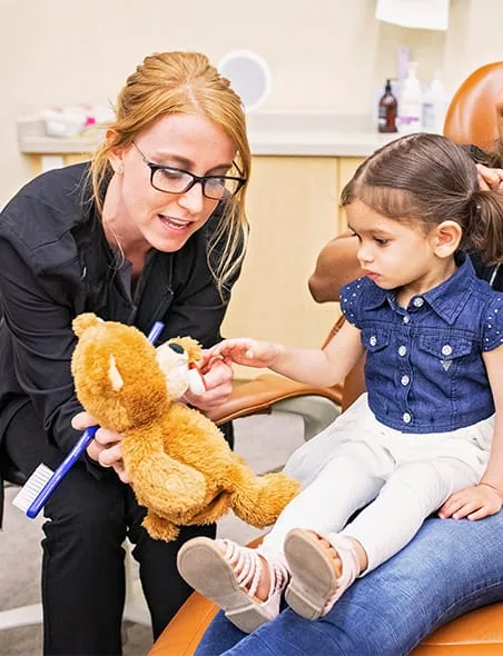 A dentist teaches a young girl about dental care with a teddy bear.