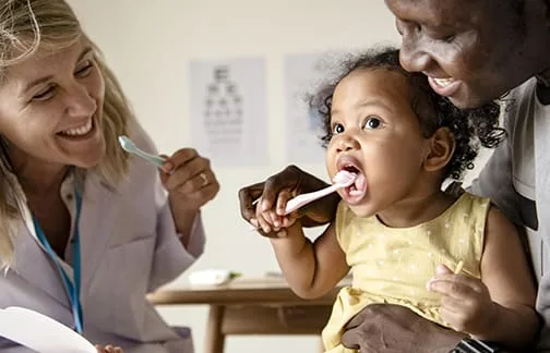 A child being assisted with brushing teeth by adults.