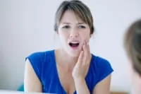 Woman in blue shirt holding cheek in discomfort
