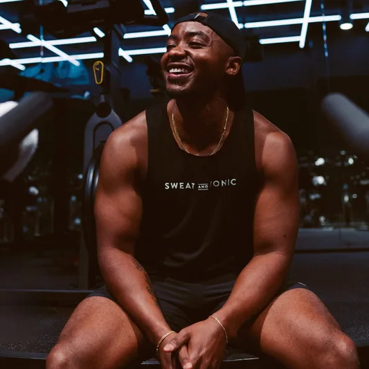 A smiling man in a gym wearing a Sweat and Tonic tank top.
