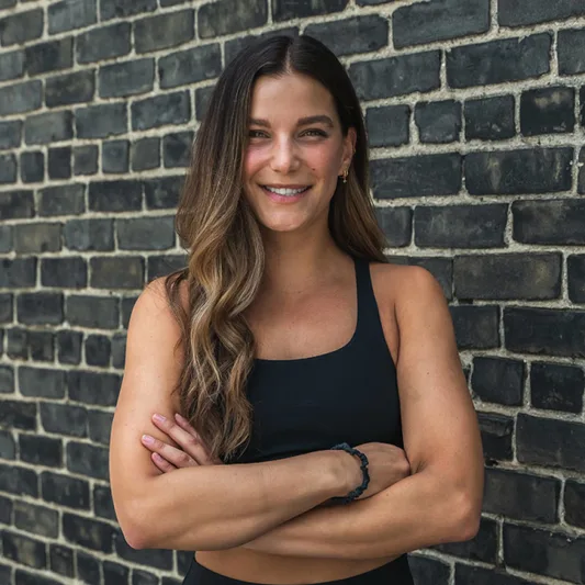 Woman with long hair in a black sports top against a brick wall.