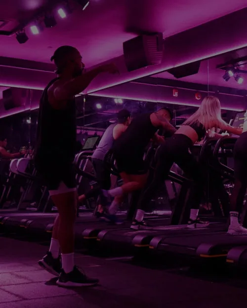 Participants in a fitness class exercising on treadmills in purple lighting.