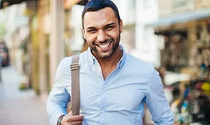 A smiling man walking on a city street with a shoulder bag.
