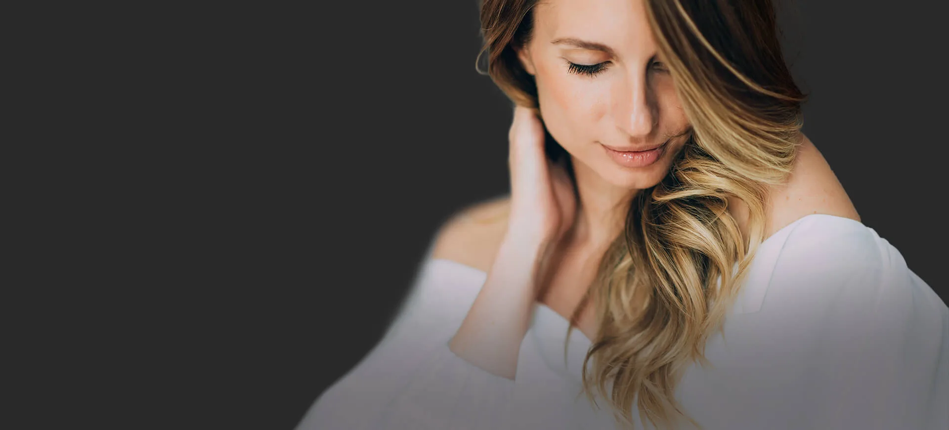 A woman in a white top touching her neck with a soft expression.